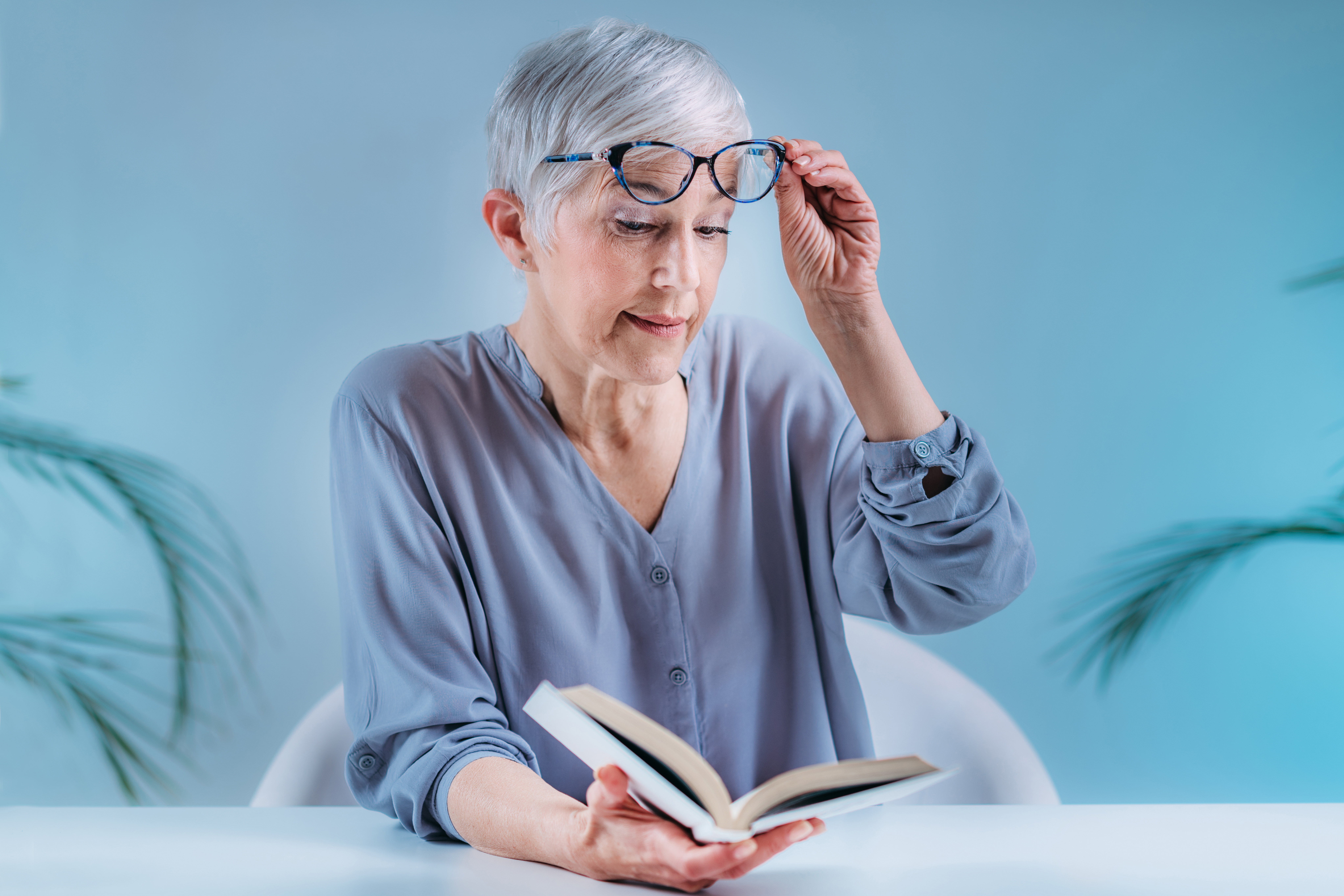 Older woman with gray hair lifts her glasses while reading a book, experiencing blurry vision from cataracts